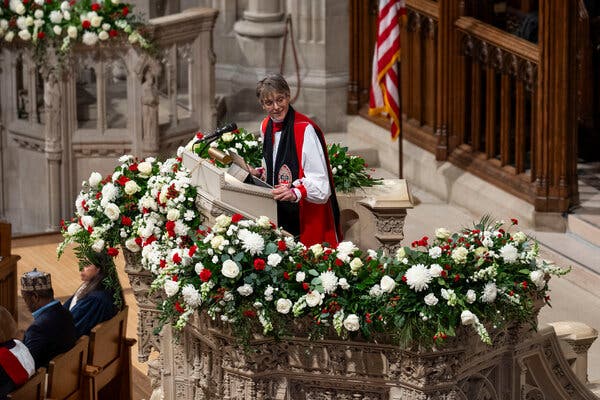 Bishop Mariann Edgar Budde, wearing a white blouse, a red tunic and a black scarf, speaks at a dais garlanded with flowers. An American flag is behind her.