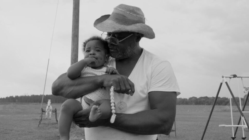 A man in a cowboy hat holds an infant on a playground.