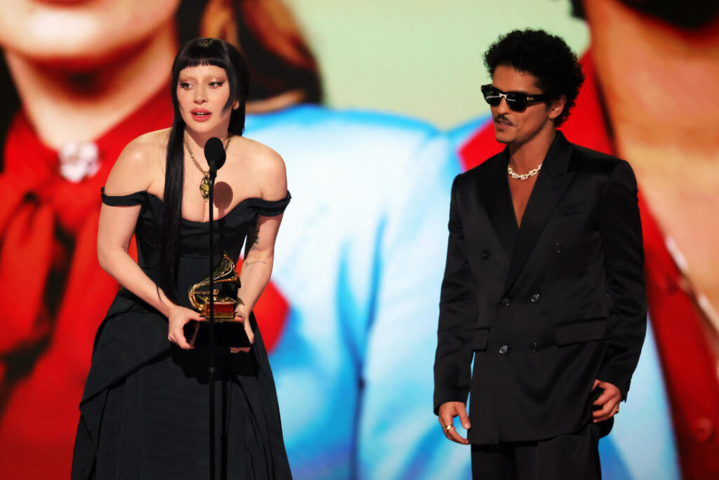 Lady Gaga holds the award at the microphone, wearing in a black gown with straps, with Bruno Mars, in dark glasses and a black suit, at her side.