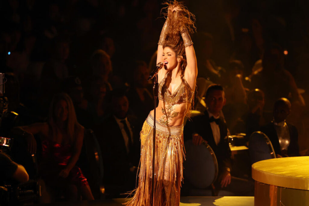 Shakira at the microphone in a fringed skirt pulling her hair above her head.