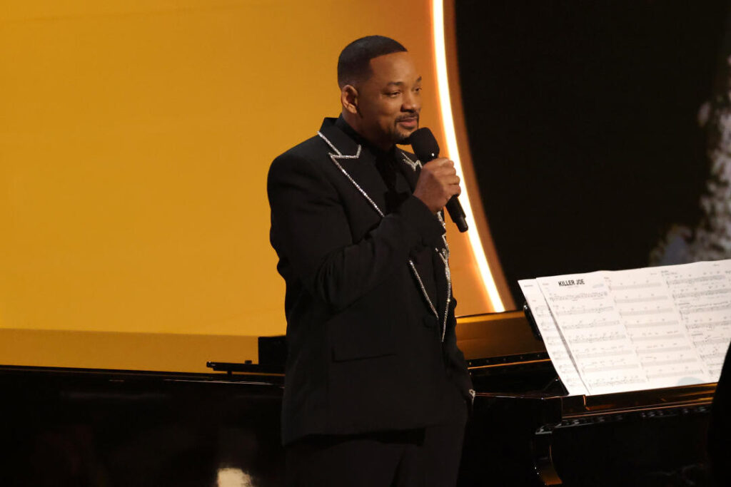 Will Smith speaks into a handheld mic as he stands next to a piano.