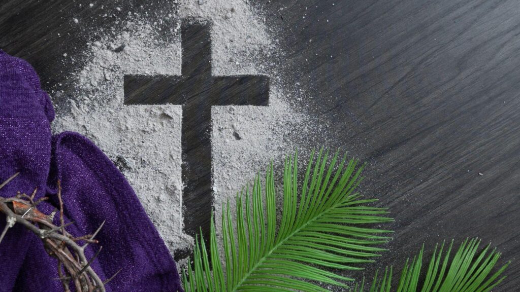 Christian lent background with cross of ashes, palm fronds, purple cloth and partial crown of thorns on a dark wood background with copy space